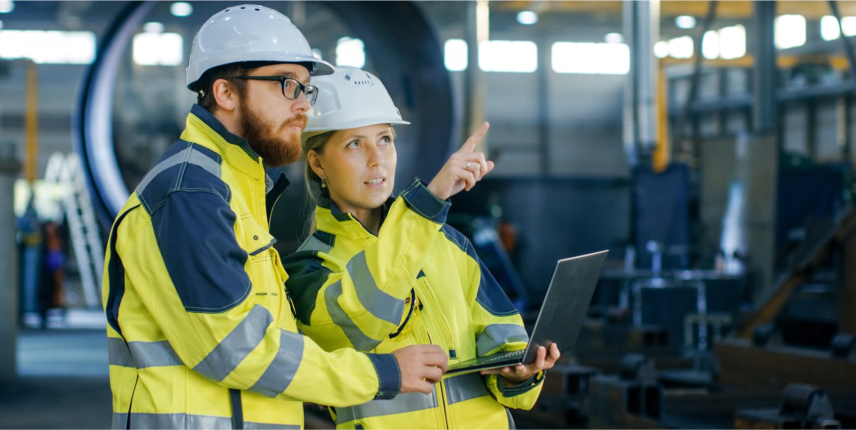 Man and woman wearing hi-vis gear in an industrial setting, holding a laptop and pointing at something out of sight.