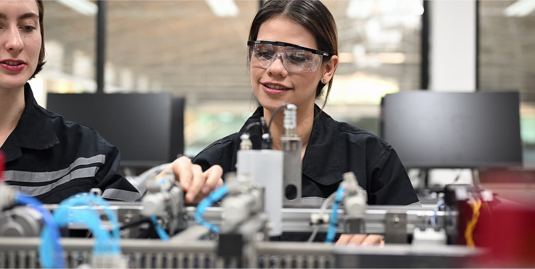 Two woman, one with safety glasses, look at some machinery.