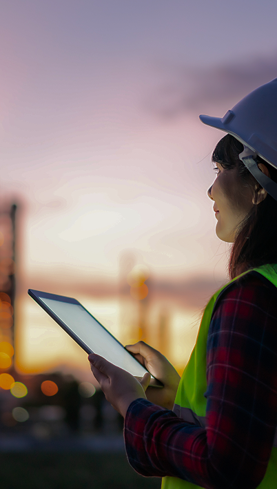 Young woman in hi-vis gear, holding a laptop and looking out towards an industrial operation.