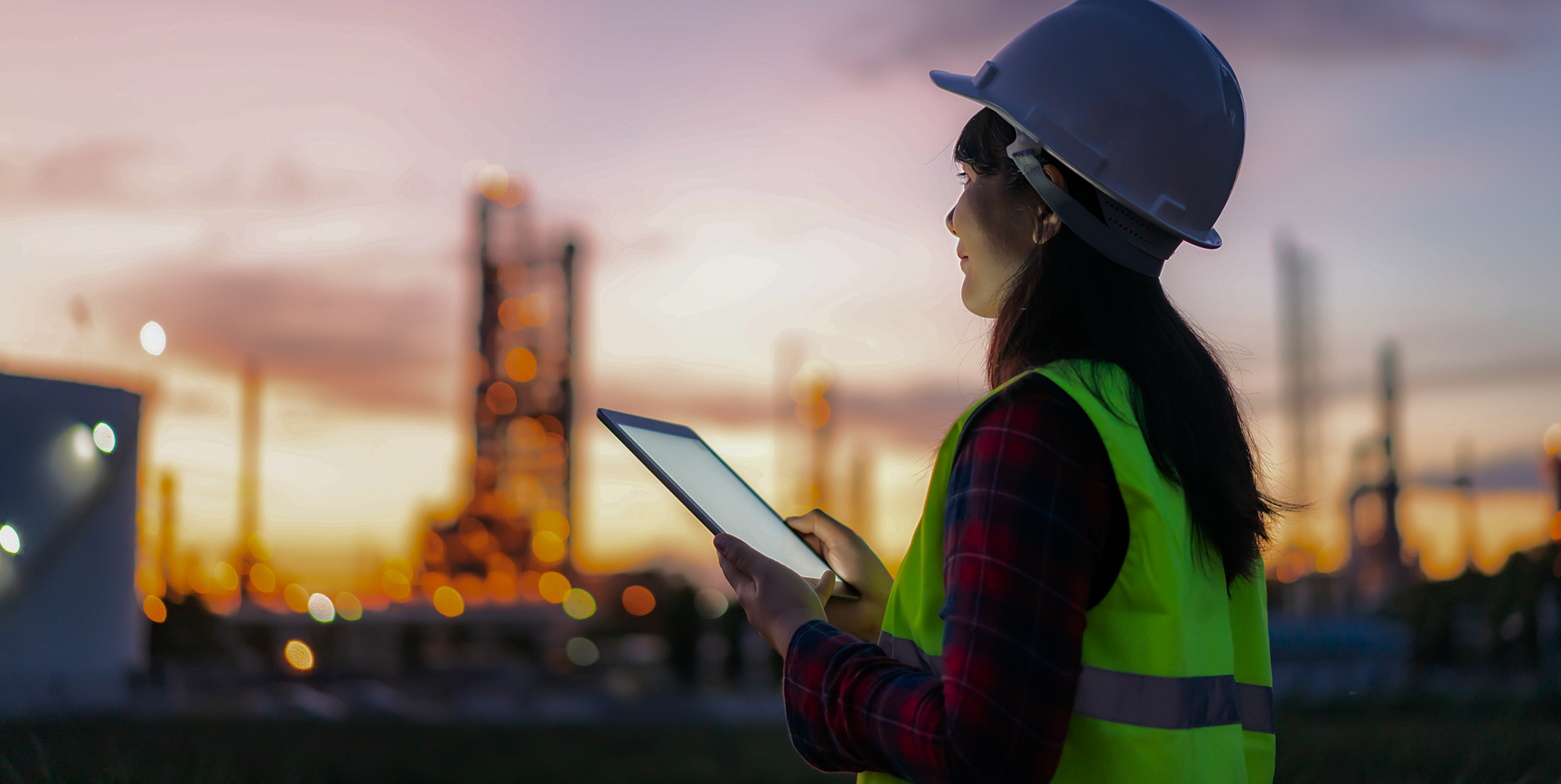 Young woman in hi-vis gear, holding a laptop and looking out towards an industrial operation.